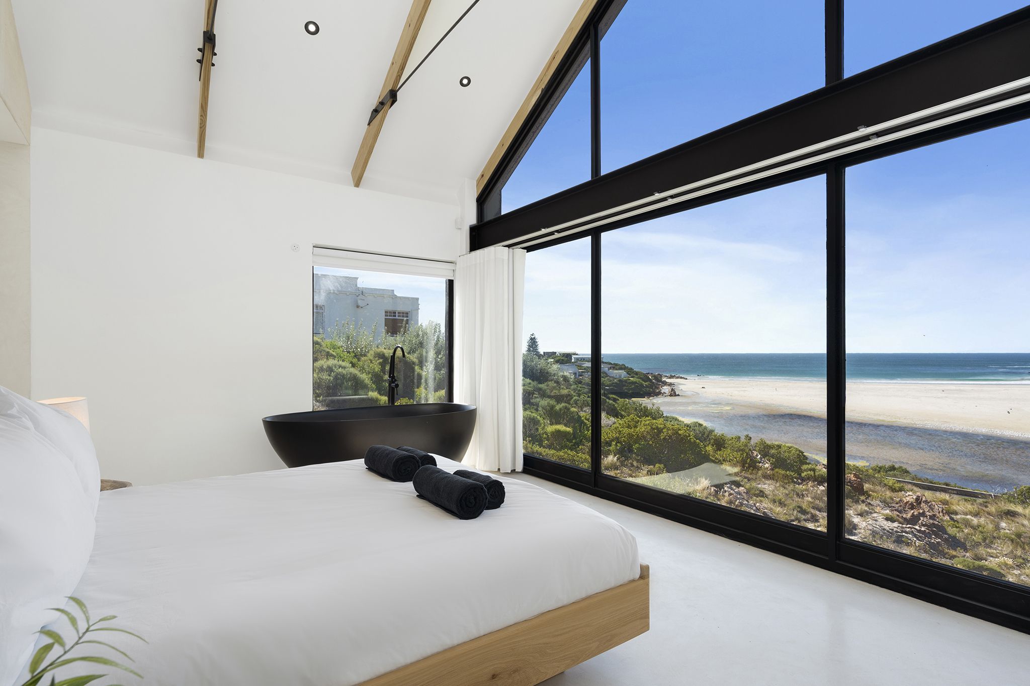A bedroom with a large window overlooking a beach, a black bathtub, and a neatly made bed.