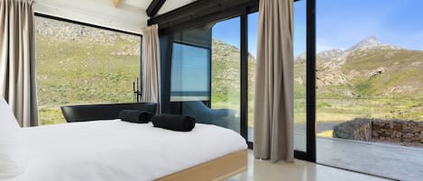 Un dormitorio moderno con una cama grande, vista a un paisaje montañoso a través de ventanales del suelo al techo, y una pared de piedra en el exterior.