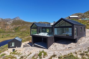 Una casa moderna de color fosc amb grans finestres de vidre i un teulat inclinat, situada en un turó rocos on s’albira un paisatge ampli i clar.