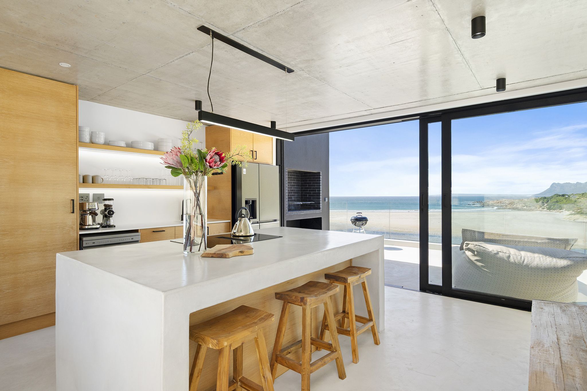 A modern kitchen with a large island, wooden stools, and a view of the beach.