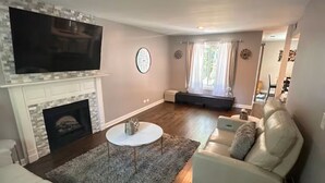 Ein Wohnzimmer mit Kamin, einem flachbildfernseher darüber, einem runden Couchtisch und einem beigefarbenen Sofa.