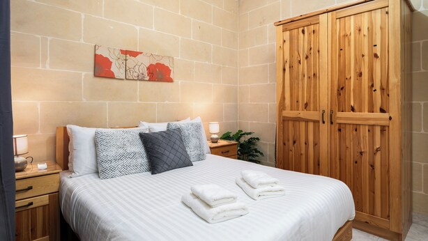A neatly made bed with white linens and two folded towels, a wooden wardrobe, and a nightstand with a lamp and a small plant. - Traditional 2BR - Terrace Country Views (Qala)
