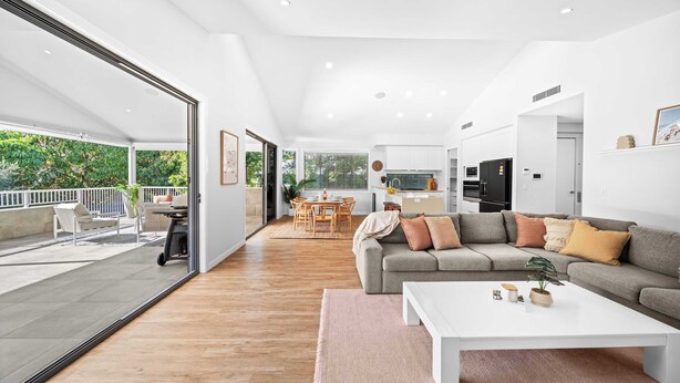 Ein geräumiger Wohnbereich mit einer großen Glasschiebetür, die zu einer Außenpatio führt, ein graues Sofa mit Kissen, ein weißer Couchtisch und eine im Hintergrund sichtbare Küchenzeile.