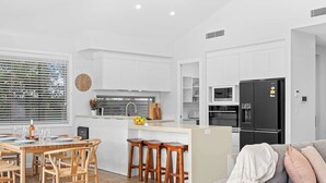 Eine moderne Küche mit weißen Schränken, ein Essbereich mit Holzsesseln und einem Tisch sowie ein graues Sofa mit einer Decke.