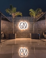 Una entrada moderna a un edificio, con un logotipo geométrico en la pared, piso de baldosas y dos palmeras.