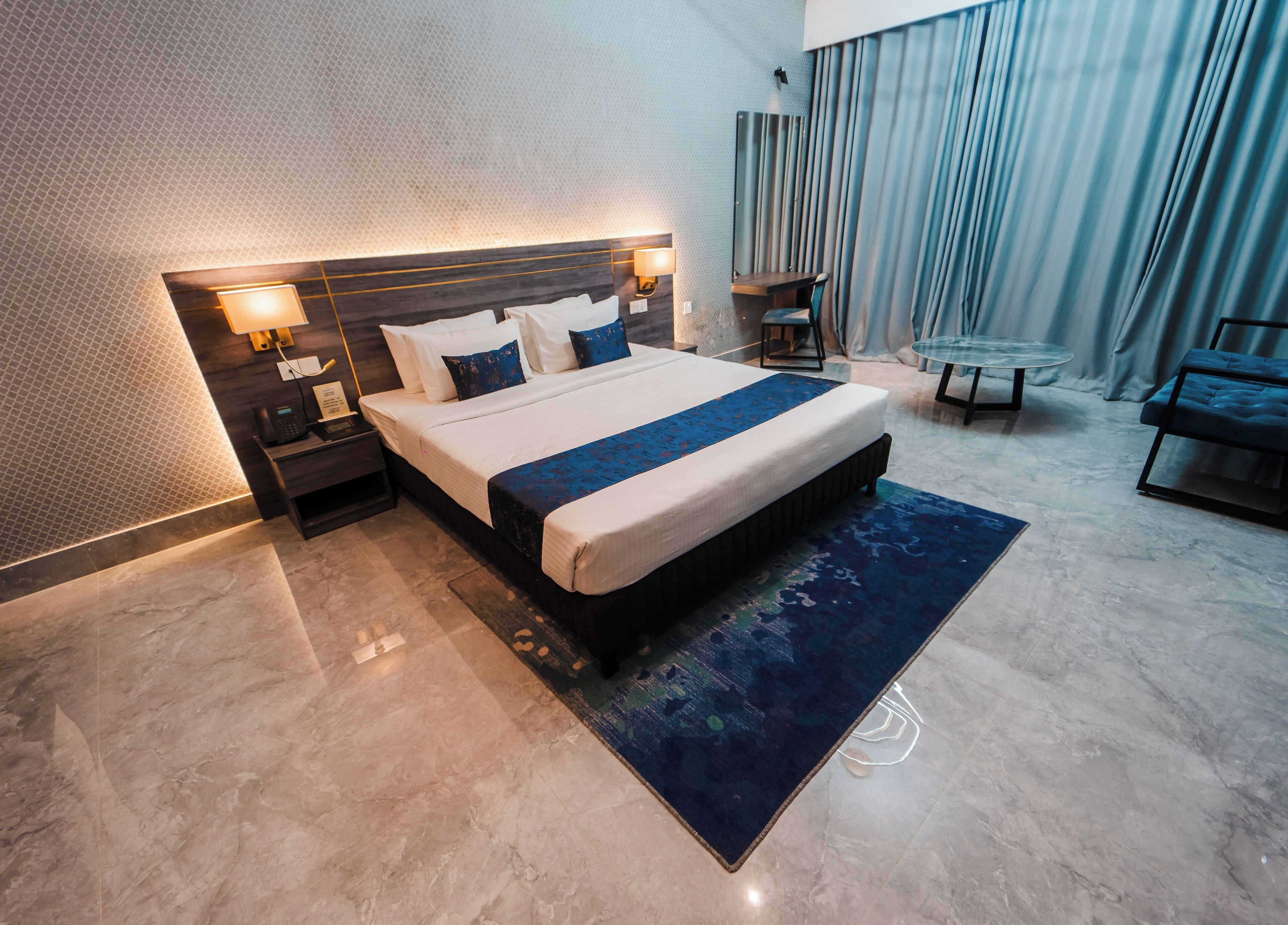 A modern hotel room with a large bed, a bedside table with a lamp, a small table with a chair, and a view of the city through large windows.