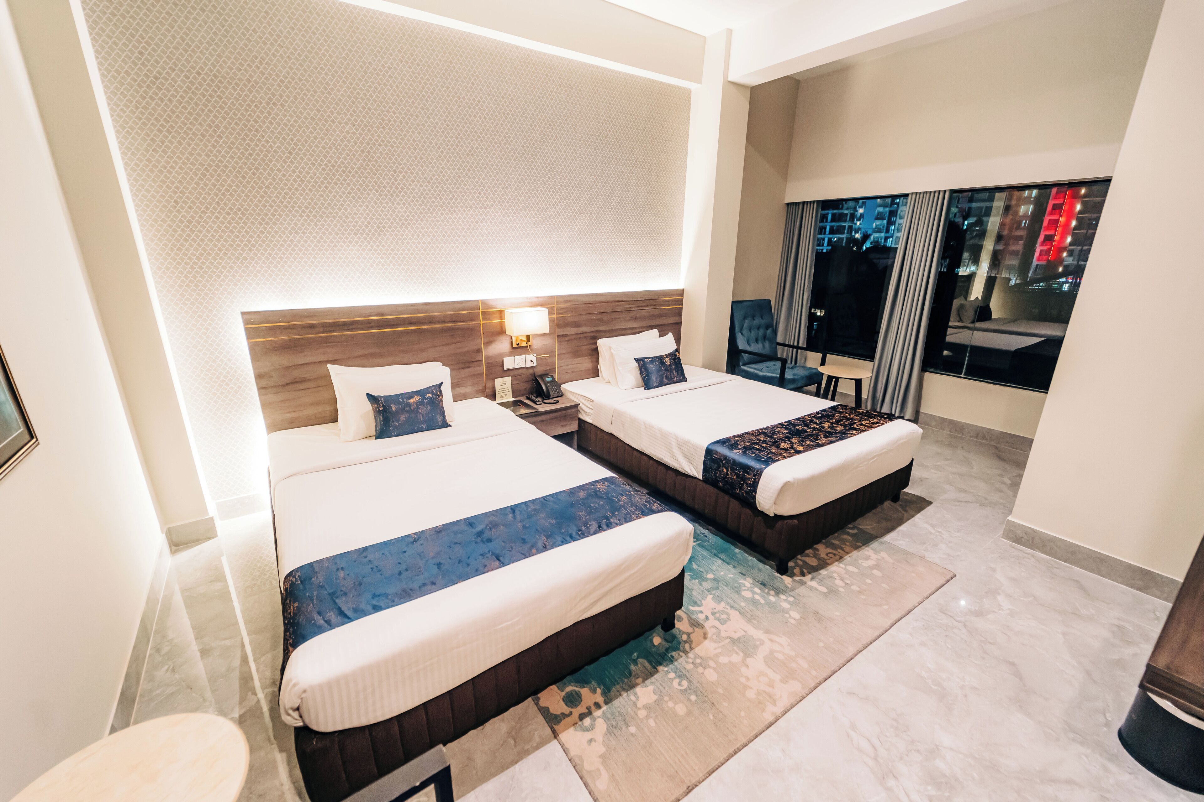 A hotel room with two beds, a large window with a view of the city, a modern headboard, and a small table.