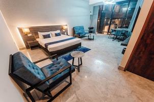 Una habitación de hotel moderna con una cama grande, una zona de estar con sofá y sillón, una mesita redonda y vistas a la ciudad a través de amplios ventanales.