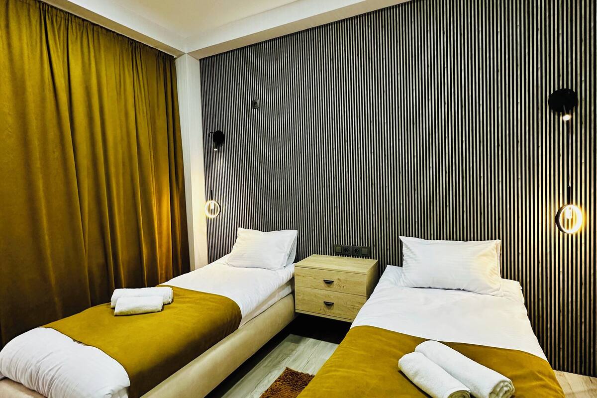 Ein Hotelzimmer mit zwei Betten, einem Nachttisch aus Holz, einer wandmontierten Leuchte und einem Vorhang.