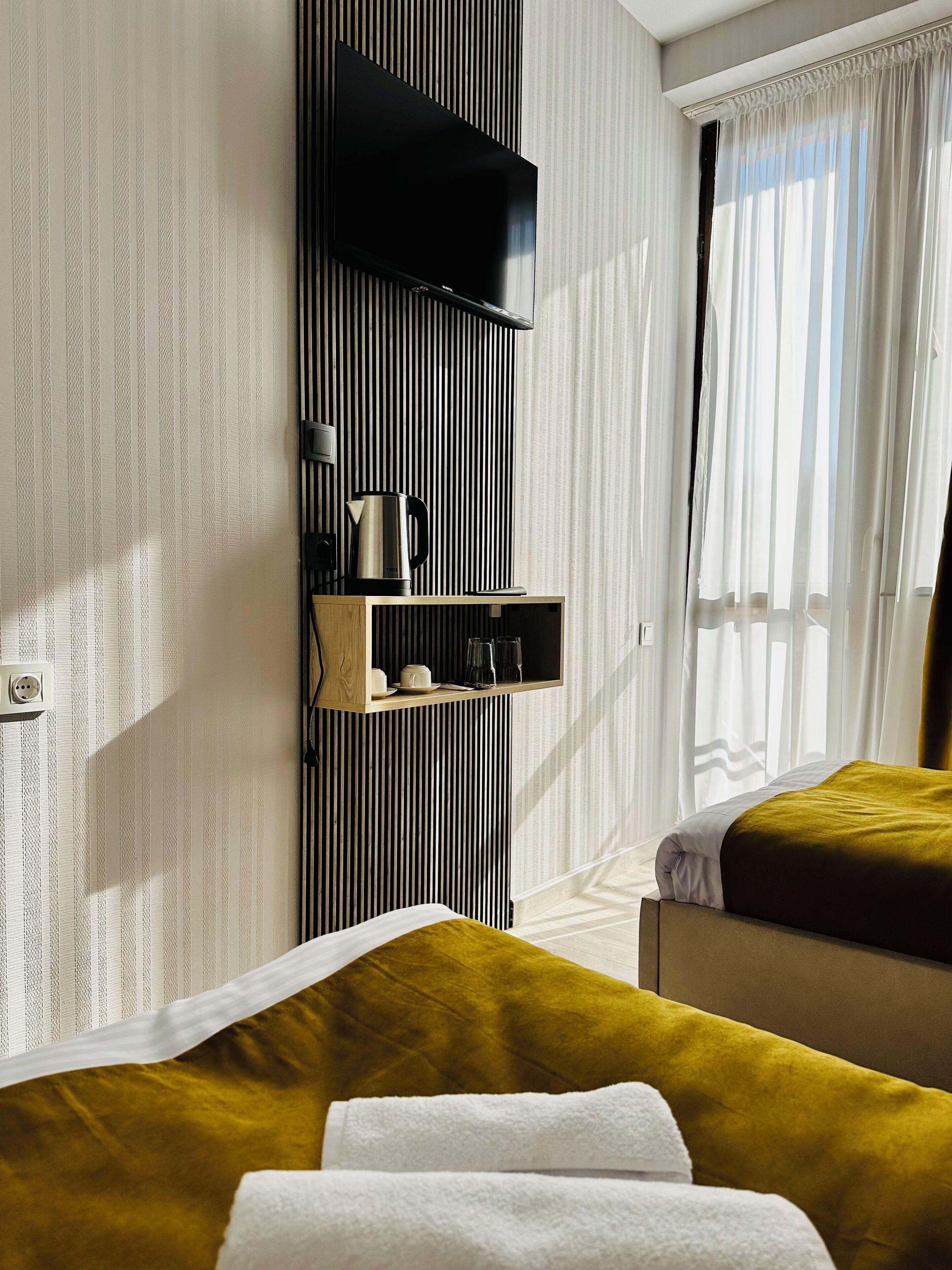A hotel room with a bed, a bedside table with a kettle and glasses, a shelf with a coffee maker, and a window with curtains.