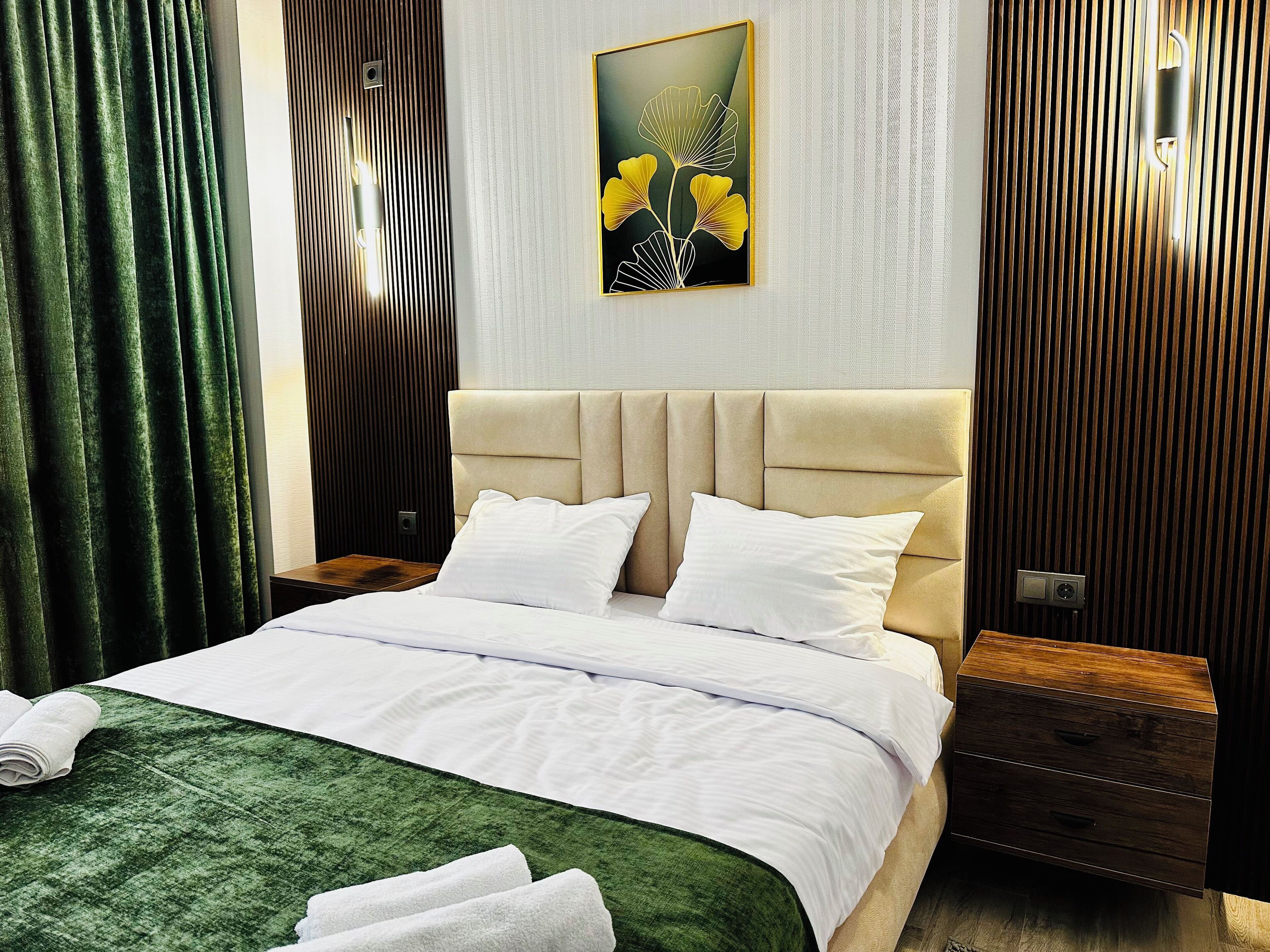 Cama bem arrumada com lençóis brancos, manta verde com textura e duas almofadas. Tem uma mesa de cabeceira de madeira com gaveta, uma luminária fixada na parede e uma obra de arte emoldurada sobre a cama.