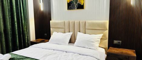 Cama bem arrumada com lençóis brancos, manta verde com textura e duas almofadas. Tem uma mesa de cabeceira de madeira com gaveta, uma luminária fixada na parede e uma obra de arte emoldurada sobre a cama.