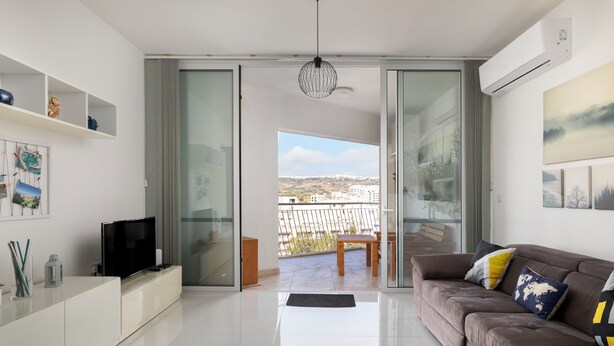 A modern living room with a flat-screen TV, a brown sofa, and a glass door leading to a balcony with a city view. - 3BR Country Retreat with Large Terrace (Zebbug)