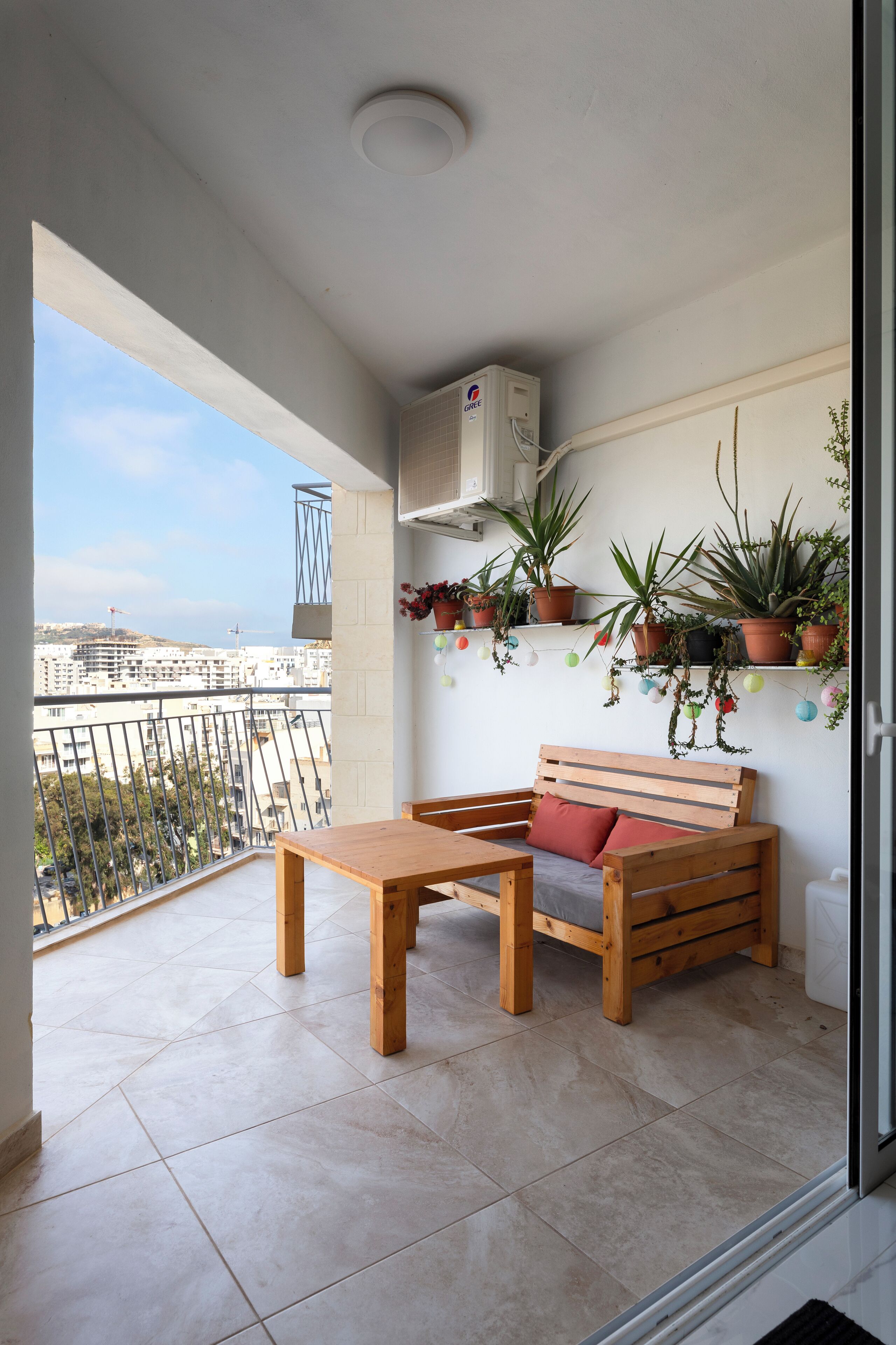 Een balkon met een houten bankje en tafel, planten in potten, en uitzicht over de stad.