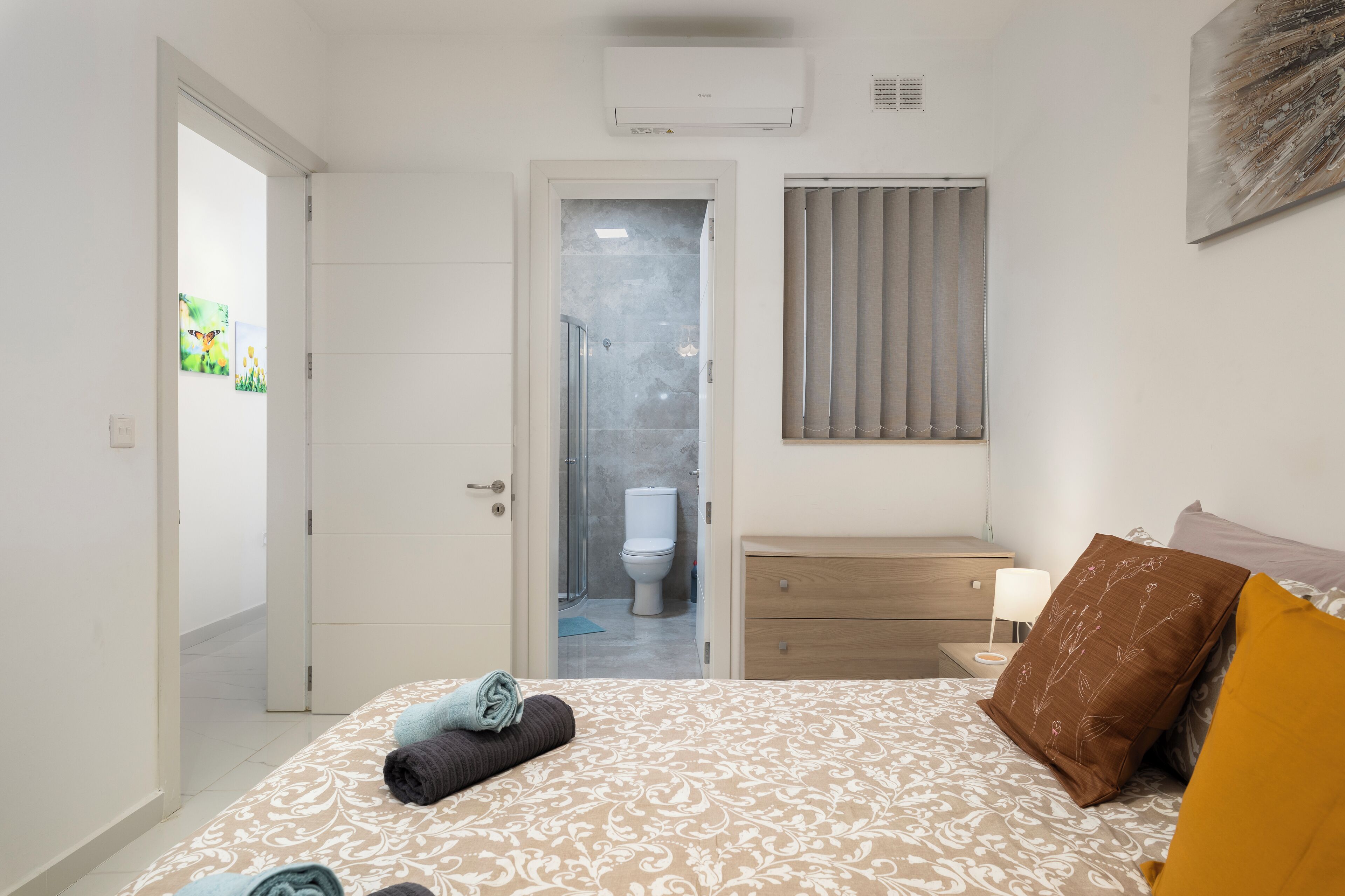A bedroom with a bed, a bedside table, a nightstand, a wall-mounted air conditioner, a window with blinds, and a door leading to a bathroom.