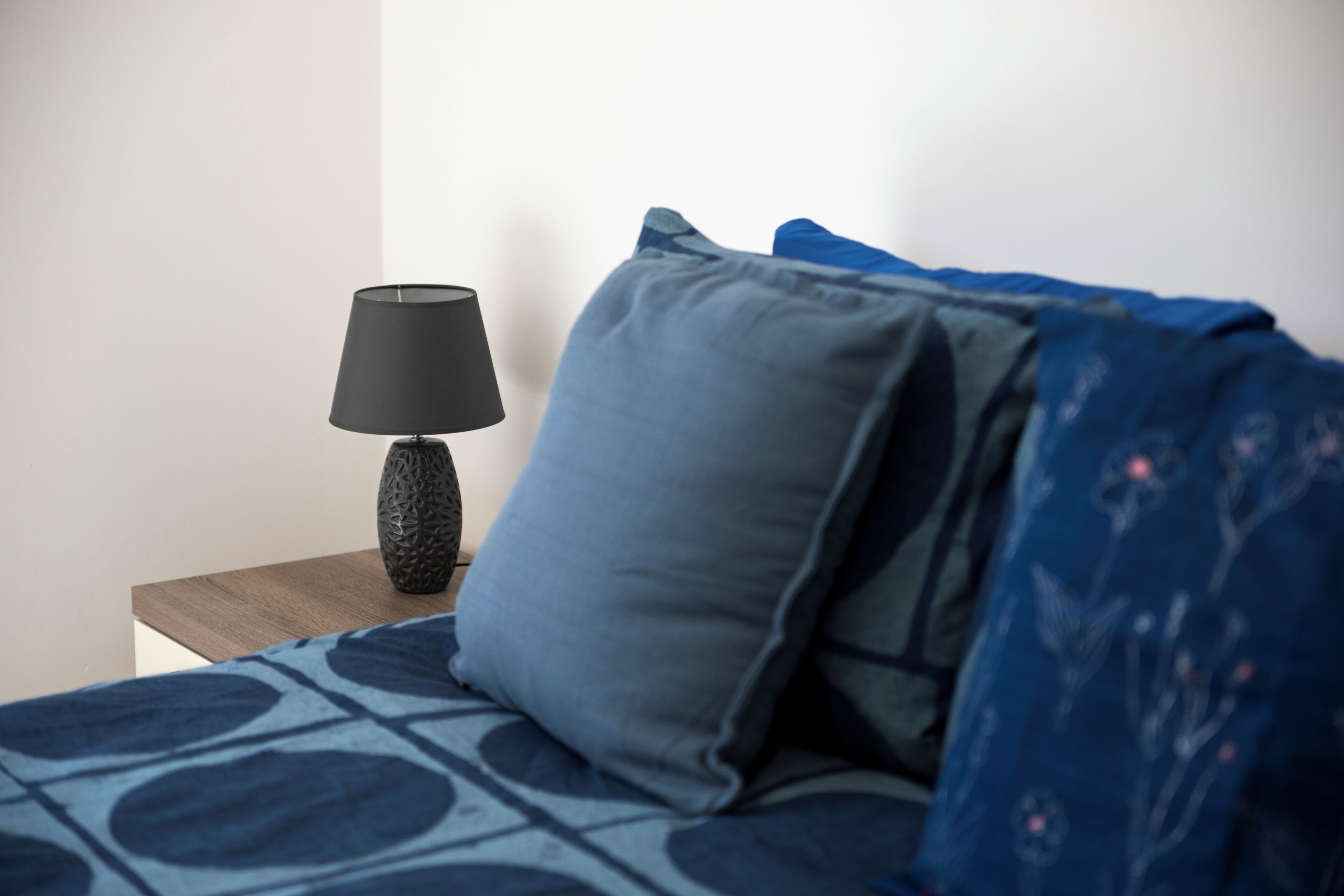A bed with a patterned blue cover, a dark blue pillow, and a black lamp on a wooden nightstand.