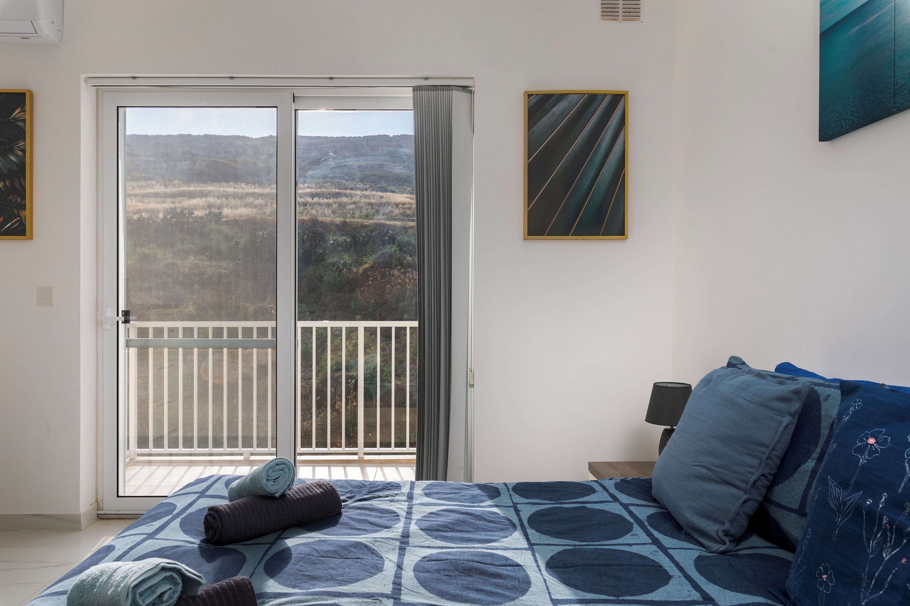 A bedroom with a bed, a nightstand, a lamp, and a view through a sliding door to an outdoor balcony.