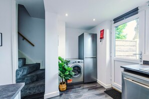 A compact kitchen with a washing machine, a stainless steel refrigerator, and a window with a view of greenery. - 2BDR Droylsden House | Free Street Parking (Manchester)