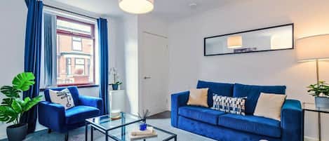Una sala moderna con un sofá azul, una mesa de centro de vidrio y vista a un edificio a través de una ventana con cortinas azules.