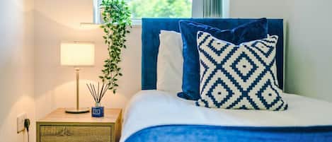 Una cama bien hecha con cabecera azul y almohadas estampadas, una mesita de noche con una lámpara y una planta, y vista al exterior a través de una ventana.
