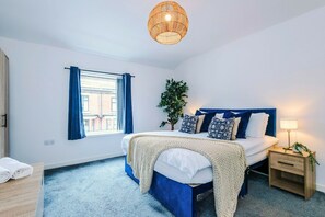 A bedroom with a bed, a bedside table, a lamp, a window with curtains, and a hanging light fixture. - 2BDR Droylsden House | Free Street Parking (Manchester)