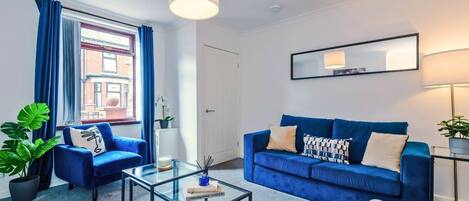 Una sala moderna con un sofá azul, una mesa de centro de vidrio y vista a un edificio a través de una ventana con cortinas azules.