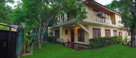 A two-story house with a balcony, surrounded by lush greenery and a well-maintained lawn.