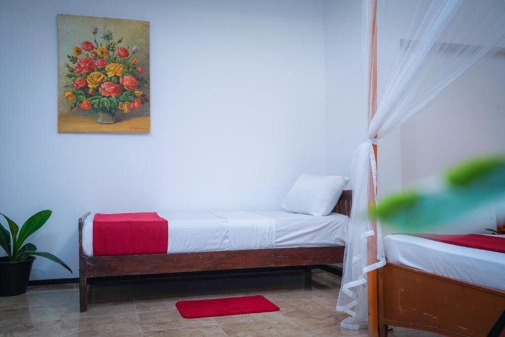 A bedroom with a wooden bed, white bedding, a red throw, a potted plant, and a painting of flowers on the wall.