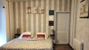Quarto com cama, mesa de cabeceira, prateleira na parede, radiador e porta.