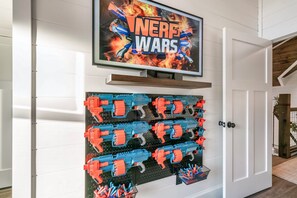 Un mur sur lequel est fixé un téléviseur diffusant l’émission « Nerf Wars », ainsi qu’un panneau perforé sur lequel sont accrochés plusieurs pistolets Nerf.