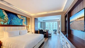 Ein modernes Hotelzimmer mit einem großen Bett, einem Schreibtisch, einem Fernseher und Blick auf die Stadt durch große Fenster.