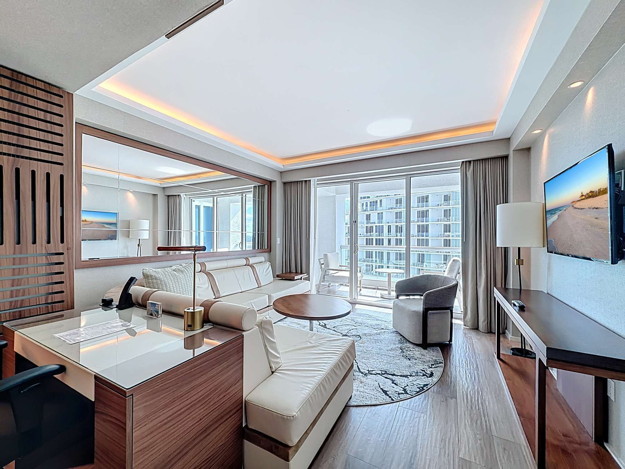 A modern hotel room with a sofa, a desk, a TV, and a balcony with a view of the city.