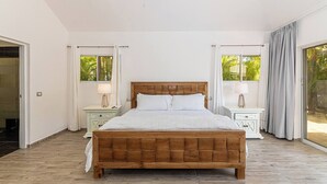 A bedroom with a wooden bed, two bedside tables with lamps, a large window with curtains, and a view of greenery outside. - Villa Ocean Dream 3BR Oasis and guesthouse (Sosúa)