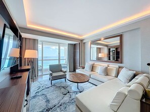 Ein modernes Hotelzimmer mit einer großen Couch, einem kleinen Sessel, einem Couchtisch und einem Fernseher. Ein großes Fenster bietet Blick auf den Ozean.
