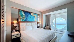 Camera d'hotel con un letto grande, un comodino, una sedia, una scrivania, un'ampia finestra vista mare e un quadro appeso al muro.