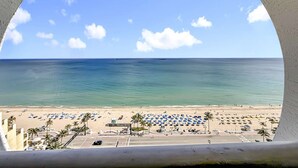 Vista di una spiaggia attraverso una finestra circolare, con spiaggia sabbiosa, ombrelloni, palme e cielo azzurro.