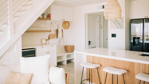 Eine moderne Küche mit weißen Schränken, einer zentralen Kochinsel mit Hockern, einer hängenden Lampenschirm aus Weidwerk und einer Treppe, die eine Ebene höher führt.
