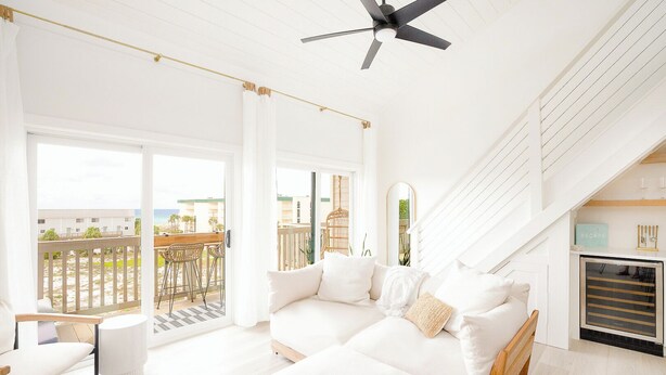 Ein geräumiges Wohnzimmer mit weißem Sofa, Deckenventilator und Blick auf den Strand durch große Fenster.