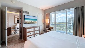 A hotel room with a large bed, a TV on the wall, a dresser, and a view of the city through large windows. - Cozy Oasis in a Luxury Hotel w/Daily Cleaning (Fort Lauderdale)