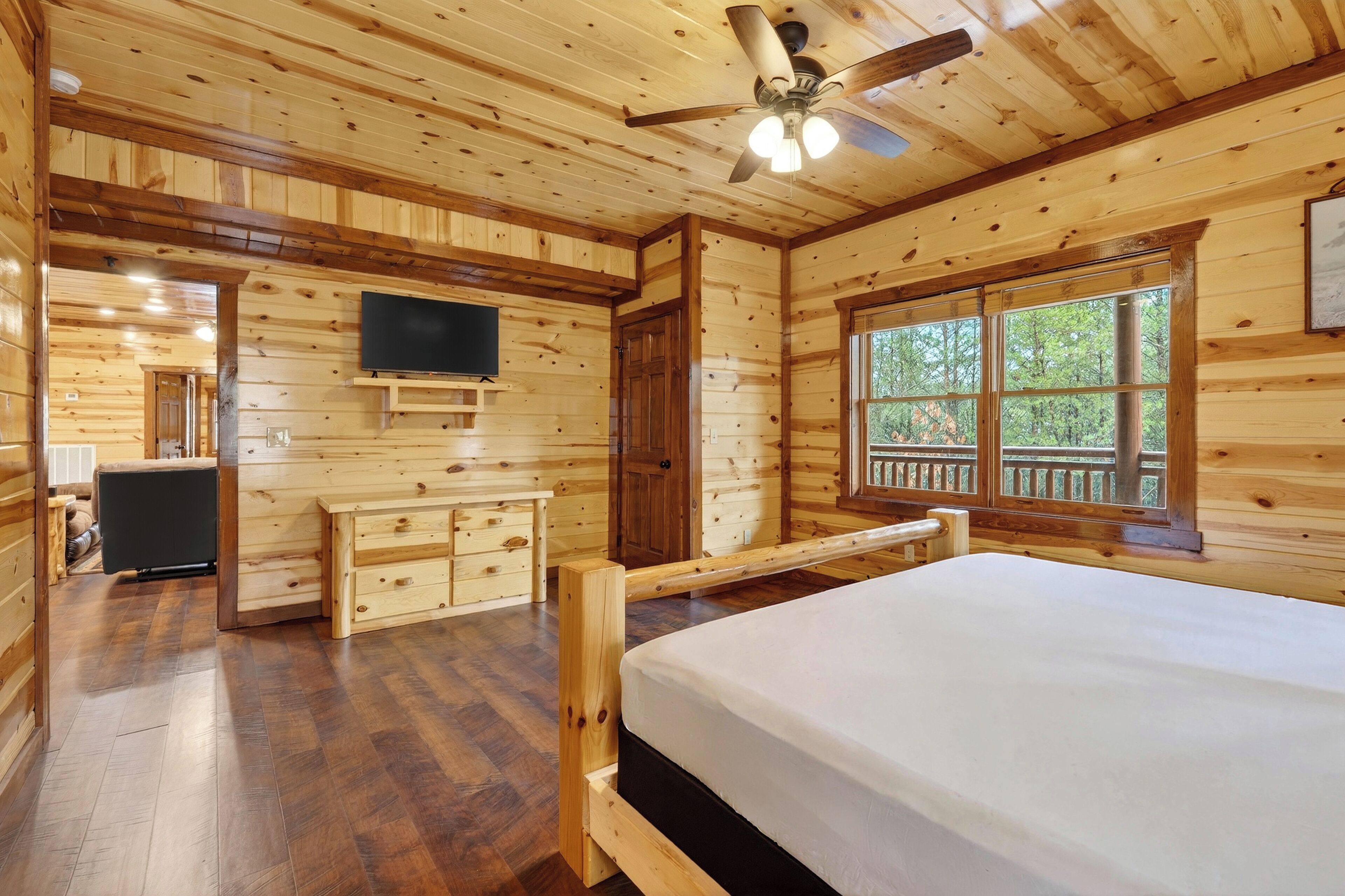 A wooden cabin bedroom with a bed, dresser, TV, and large windows.