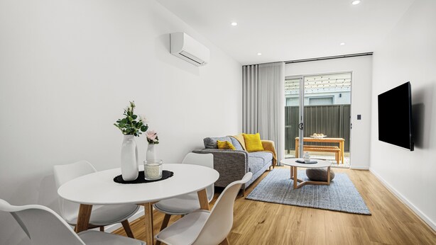 A modern living room with a dining area, a grey sofa with yellow pillows, a flat-screen TV mounted on the wall, and a large window with curtains. - Relax by the Beach, Close to Everything - Minutes from City & Airport! (Flinders Park)