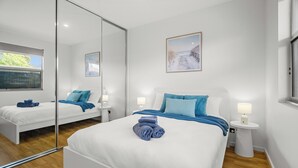 A modern hotel room with a large bed, a bedside table with a lamp, a mirror, and a window with blinds. - Relax by the Beach, Close to Everything - Minutes from City & Airport! (Flinders Park)