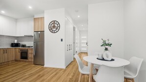 A modern kitchen with white cabinets, a stainless steel refrigerator, and a dining area with white chairs and a table. - Relax by the Beach, Close to Everything - Minutes from City & Airport! (Flinders Park)