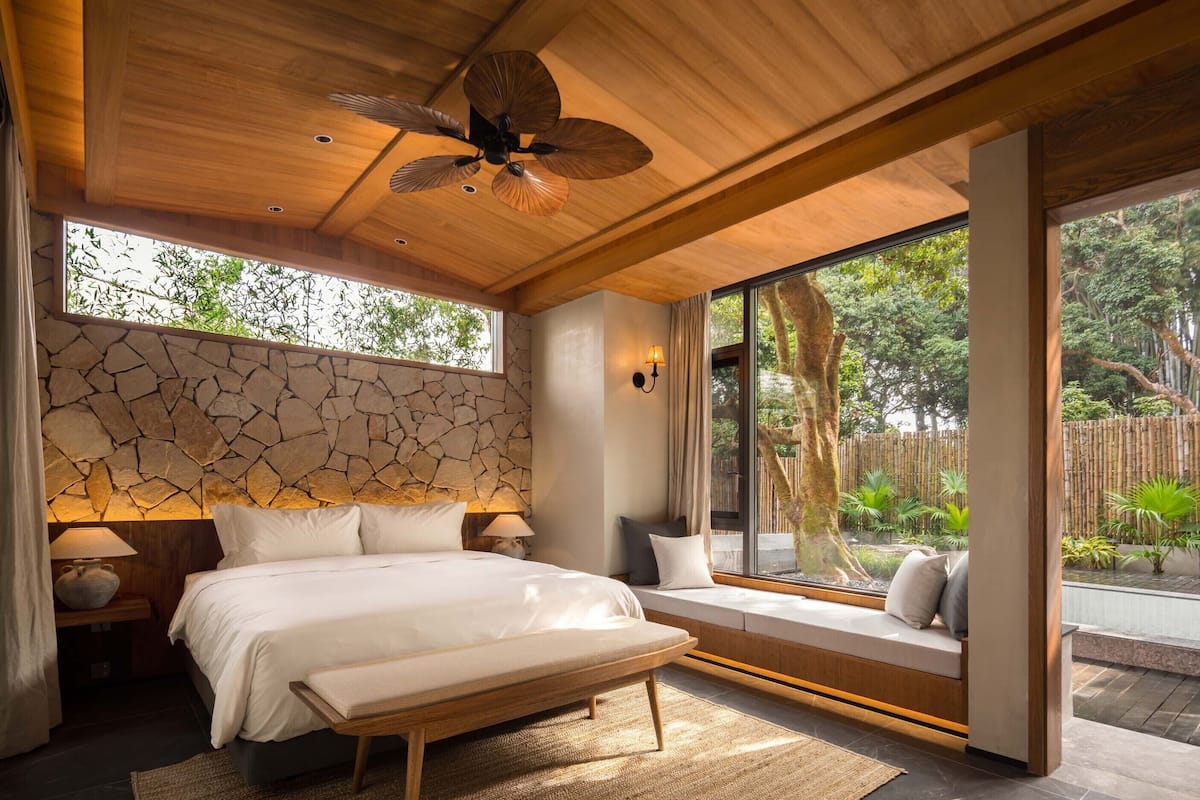 Un dormitorio con pared de piedra, una cama, un banco, ventilador de techo y ventanas amplias con vistas a un jardín.