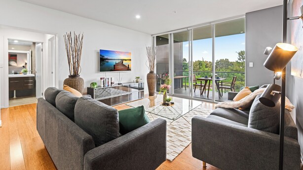 A modern living room with a sofa, a coffee table, and a TV. There is a balcony with a view of greenery. - Adelaide Parkside Escape | 2BR + Gym + Parking (Adelaide)