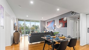A modern living space with a dining area, a sofa, and a large window. - Adelaide Parkside Escape | 2BR + Gym + Parking (Adelaide)