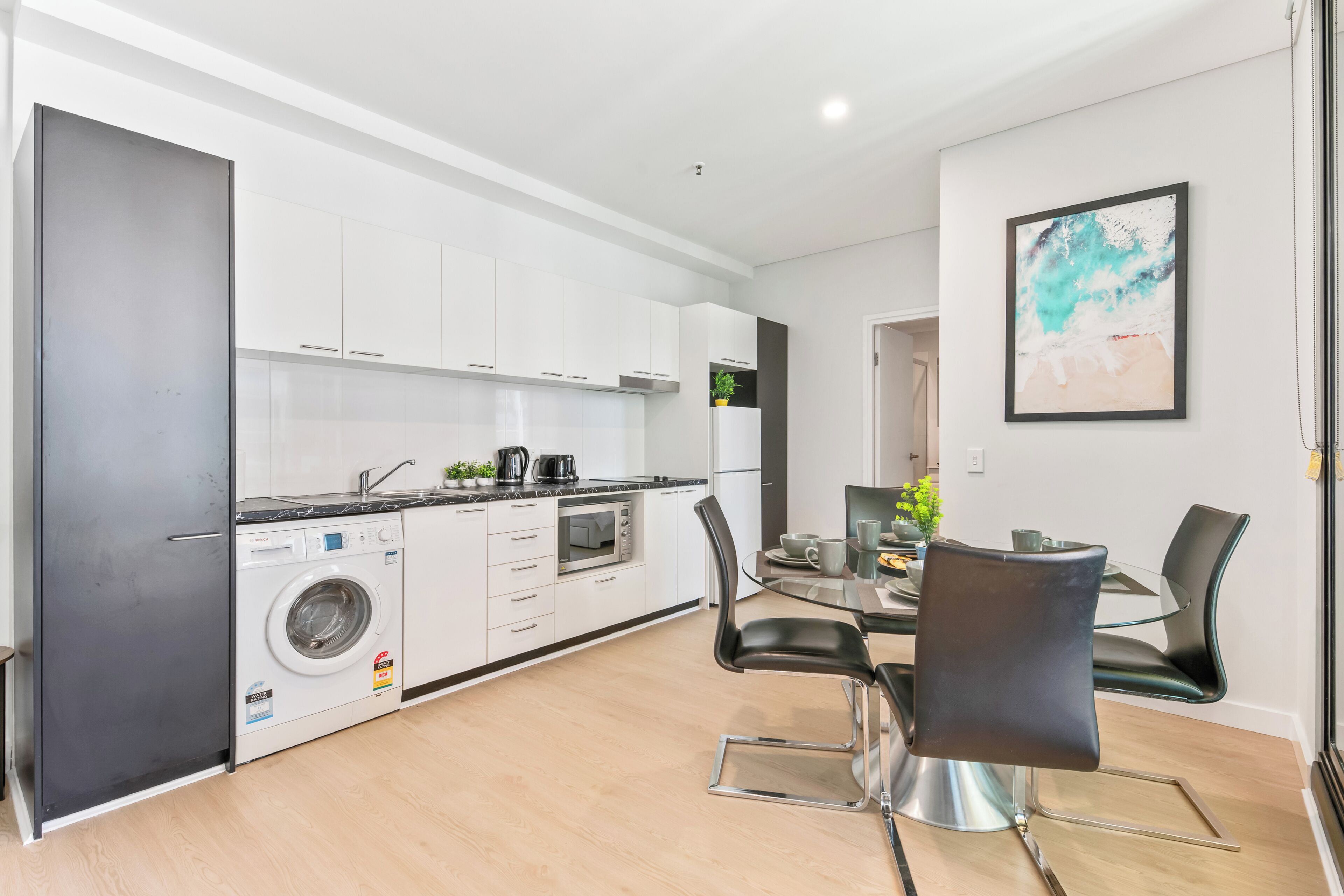 A modern kitchen with white cabinets, a built-in washing machine, and a dining area with a glass table and black chairs.