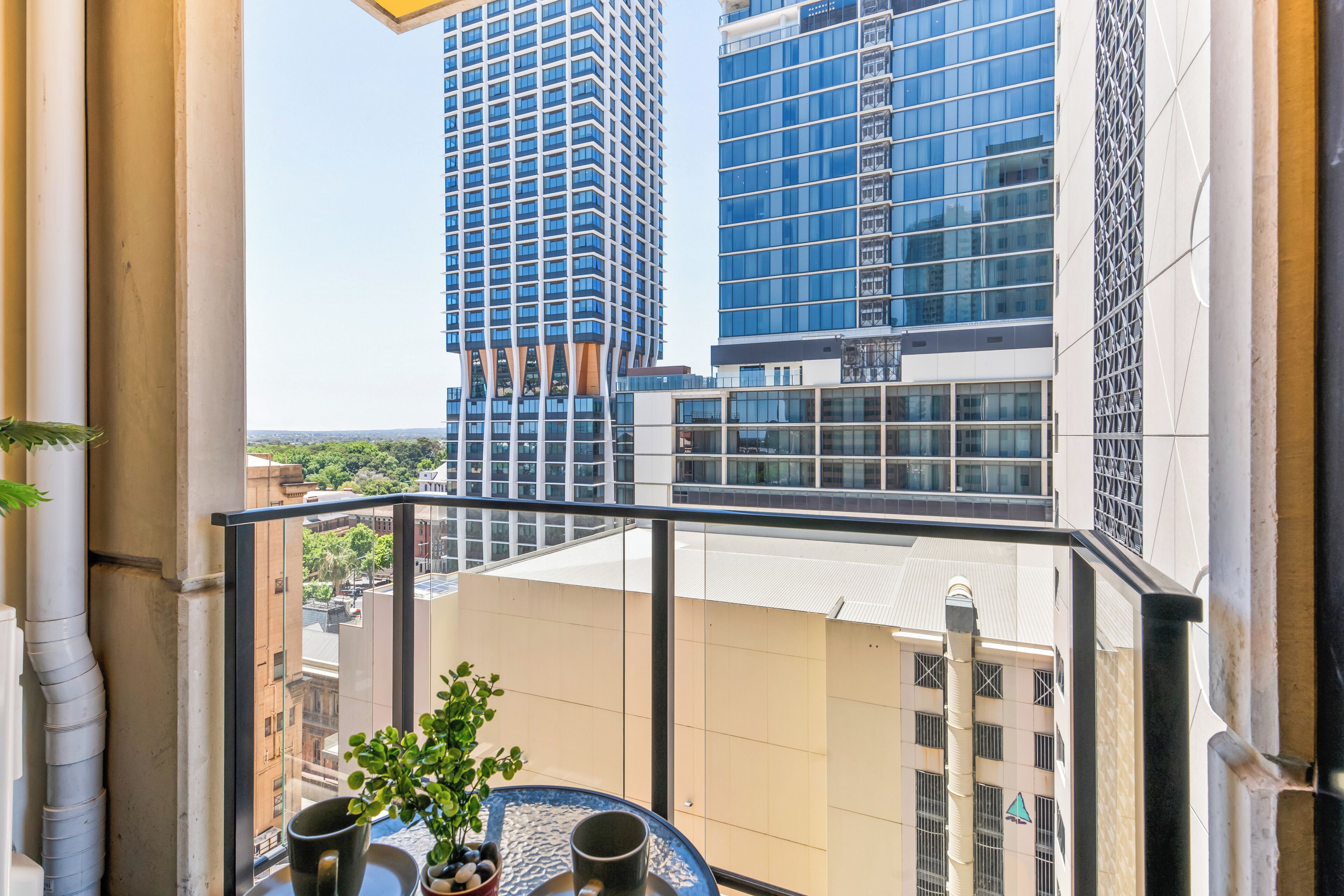 A balcony with a view of tall buildings, a potted plant, and a table with a mug.
