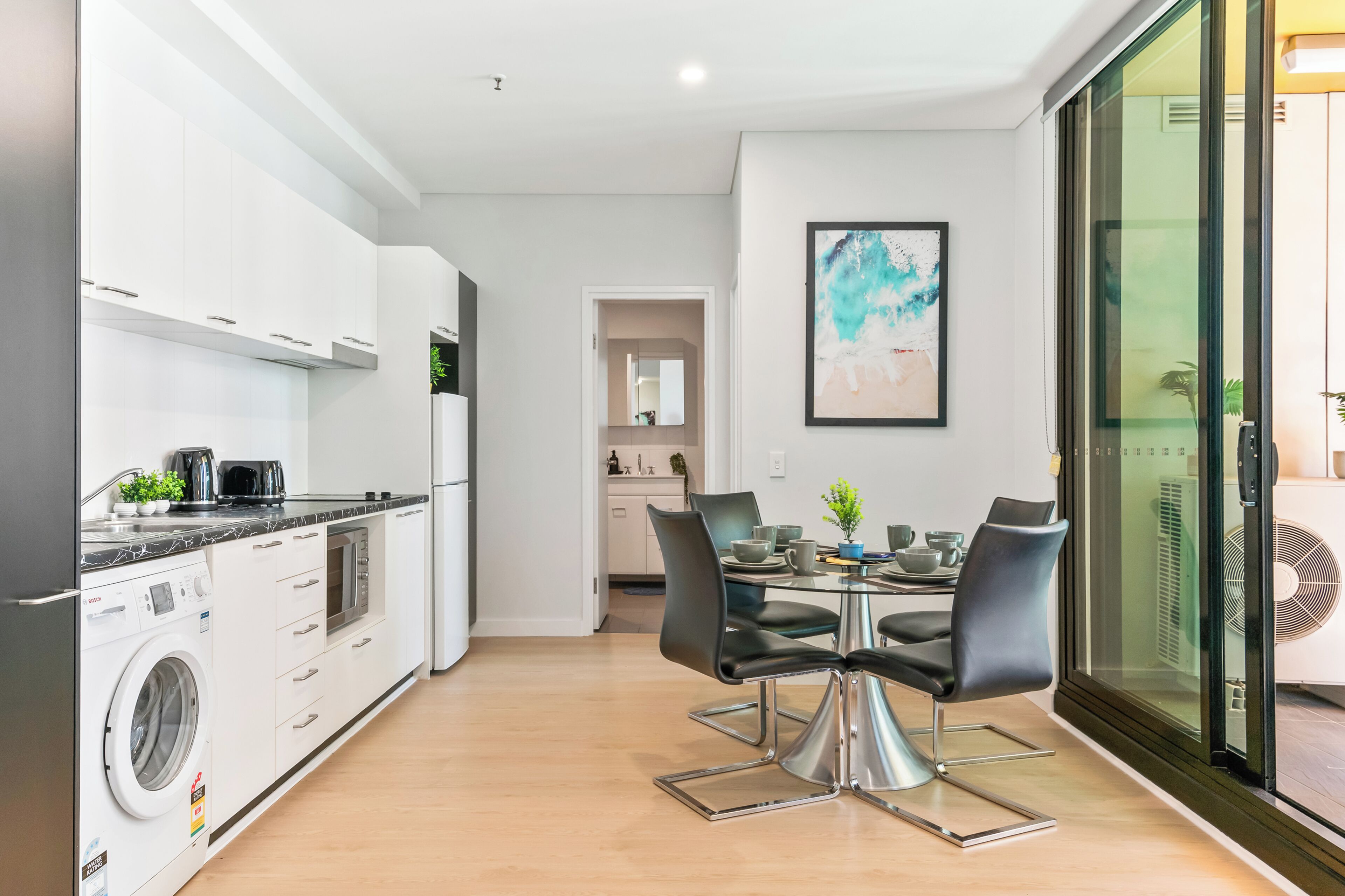 Une cuisine moderne dotée d’appareils électroménagers intégrés, un coin repas avec une table en verre et des chaises noires, et une vue à travers une porte vitrée.