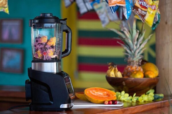 A blender with fruits inside, a wooden cutting board, and a bowl of fruits on a table. - Open Gate Hostel Hawaii (Pāhoa)
